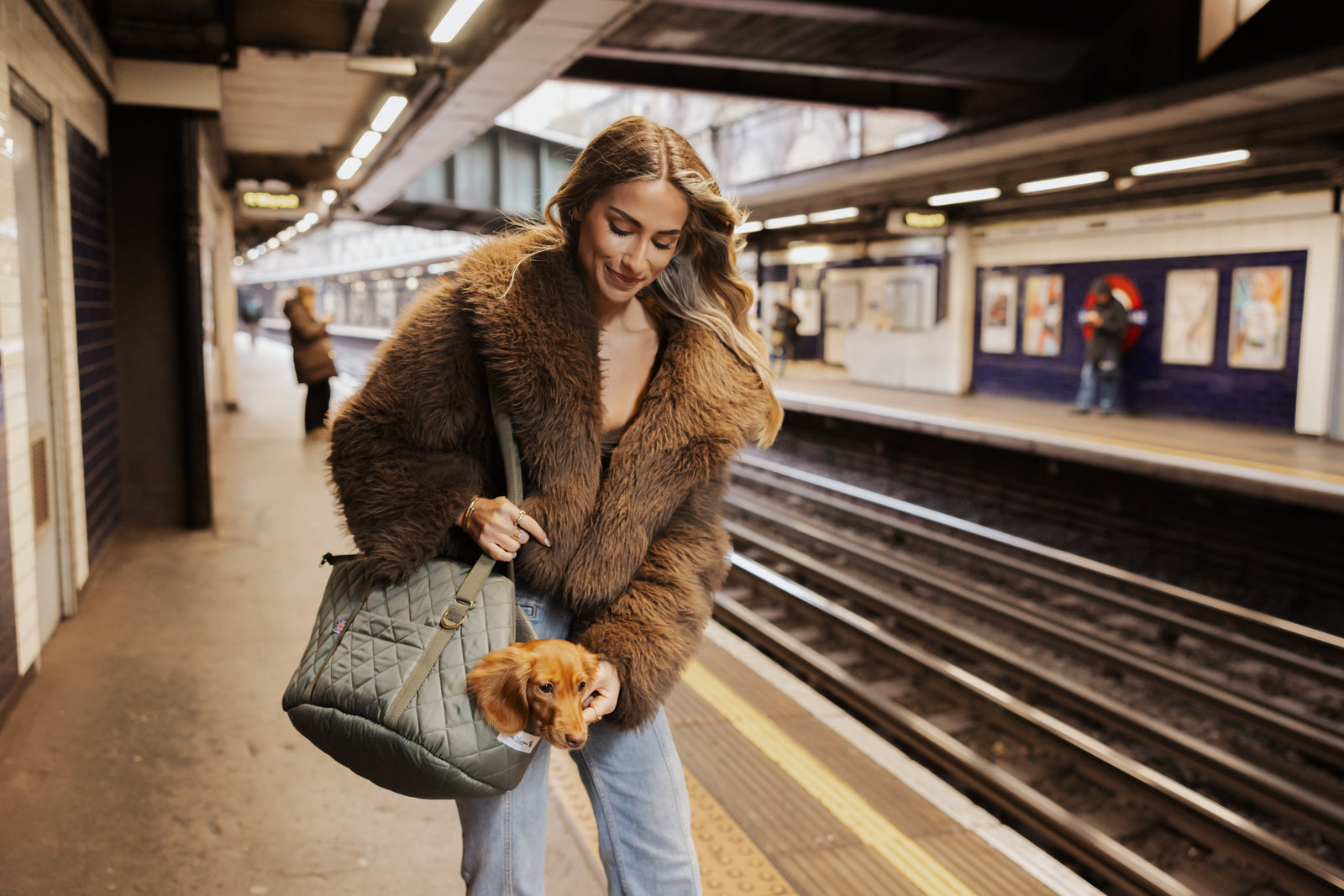 Woman in a fur coat holding a dog in a  Teddy Maximus bag on a train platform